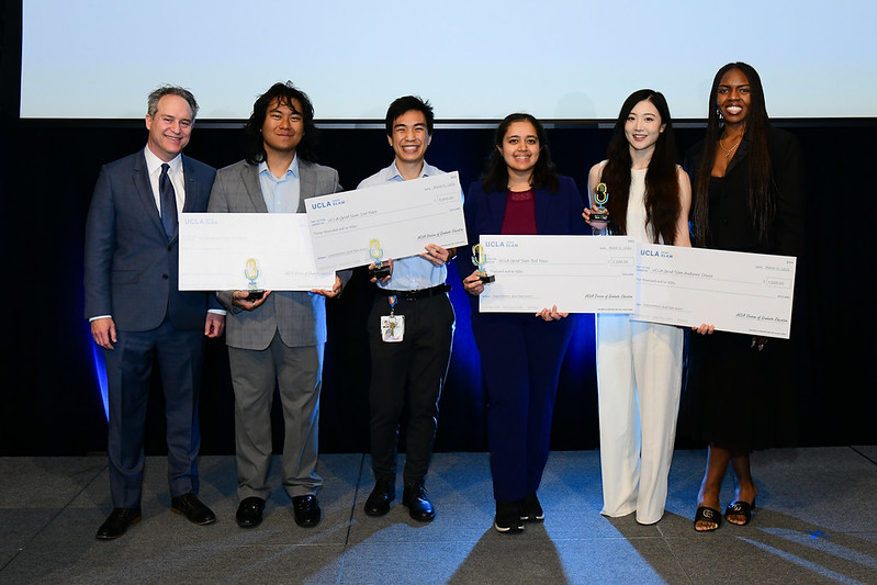 Group photo of the 2026 UCLA Grad Slam winners standing on stage with oversized checks and trophies alongside Dean and Vice Provost Brian Kite and GSA President Naomi Hammonds. From left to right: Brian Kite; first-place winner Christian Gerard Capuno; second-place winner Noah Yan; third-place winner Shreya Kordale; audience choice winner Jiaao Liu; and Naomi Hammonds. Photo by Rich Schmidt.