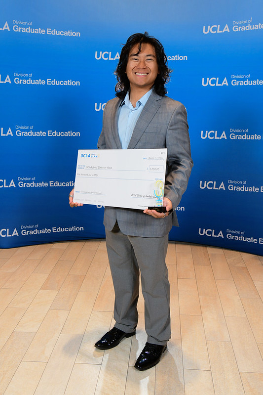 UCLA graduate student Christian Capuano smiles while holding an oversized check and trophy after winning the 2026 Grad Slam, standing before a UCLA Division of Graduate Education backdrop—capturing academic excellence, research impact, and student achievement.