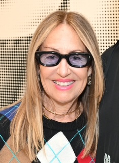 Close-up portrait of Alexandra Serros smiling at the camera, wearing dark cat-eye glasses, a black sweater with a tan and blue argyle pattern, layered gold necklaces, and long straight blonde hair against a patterned  backdrop.